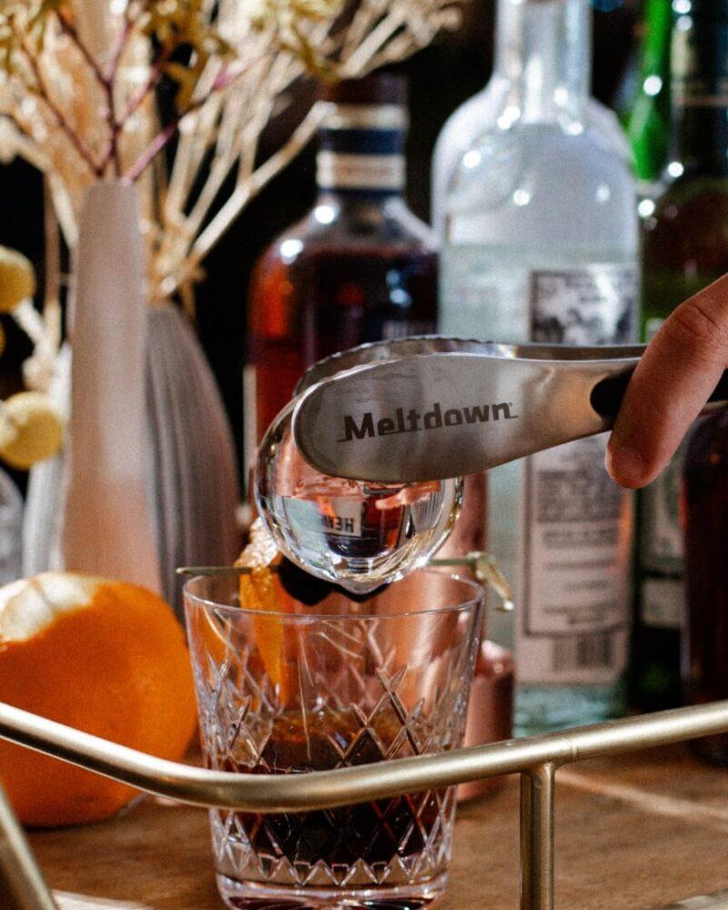 Meltdown steel tongs placing ice sphere in Negroni glass on bar cart