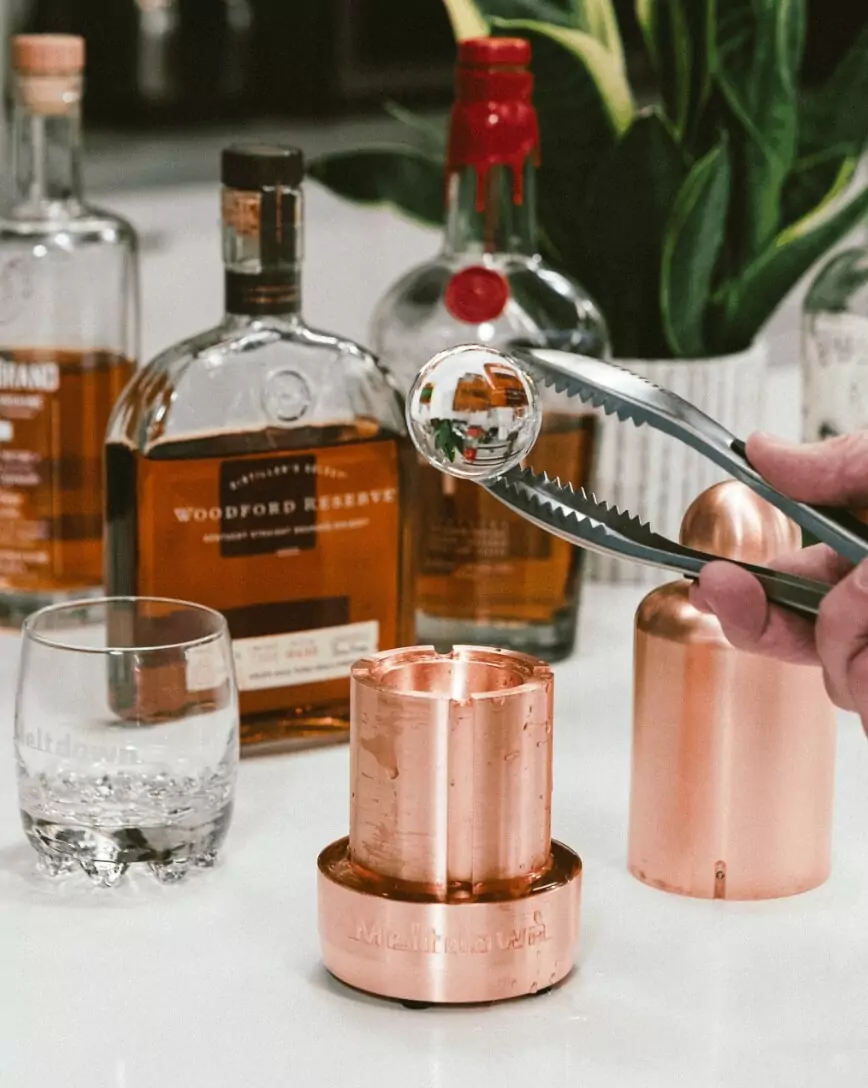 Meltdown steel tongs holding ice sphere before bottle of Woodford Reserve and Meltdown Mini Mogul