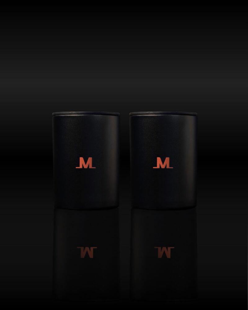 Two black mugs featuring a red M logo, inspired by the Meltdown Mogul Ice Press, sit side by side on a dark surface, their reflections shimmering below—evoking the premium quality of the Meltdown Mogul Ice Press.