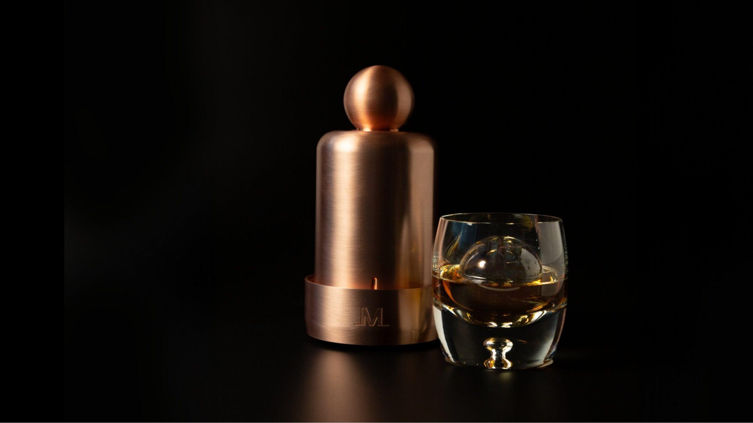 A copper-colored Meltdown ice ball press stands next to a glass of whiskey containing a large, perfectly round ice sphere, set against a black background.