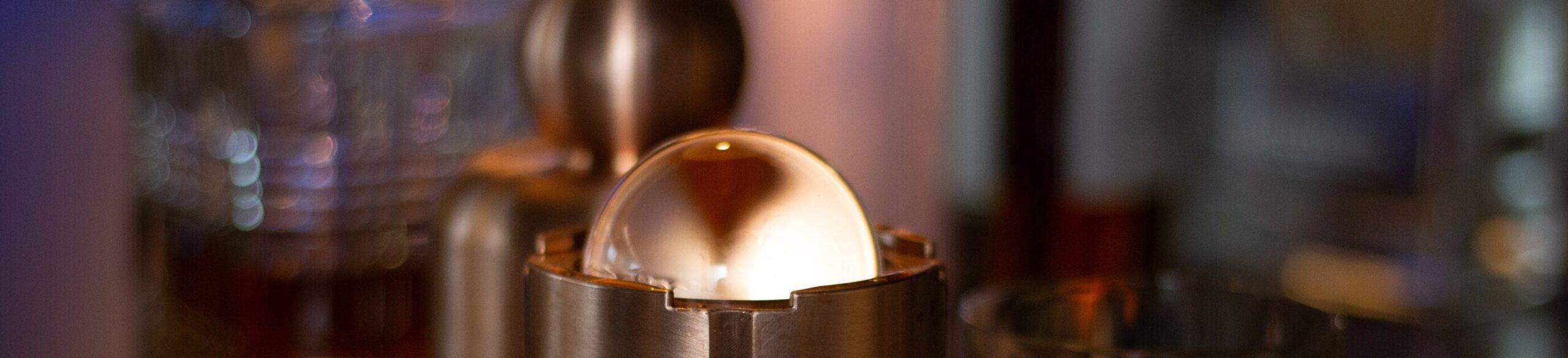 A close-up image of a clear ice sphere resting in a metallic holder, with blurred glassware and bottles in the background. Warm lighting highlights the smooth surface of the ice sphere.