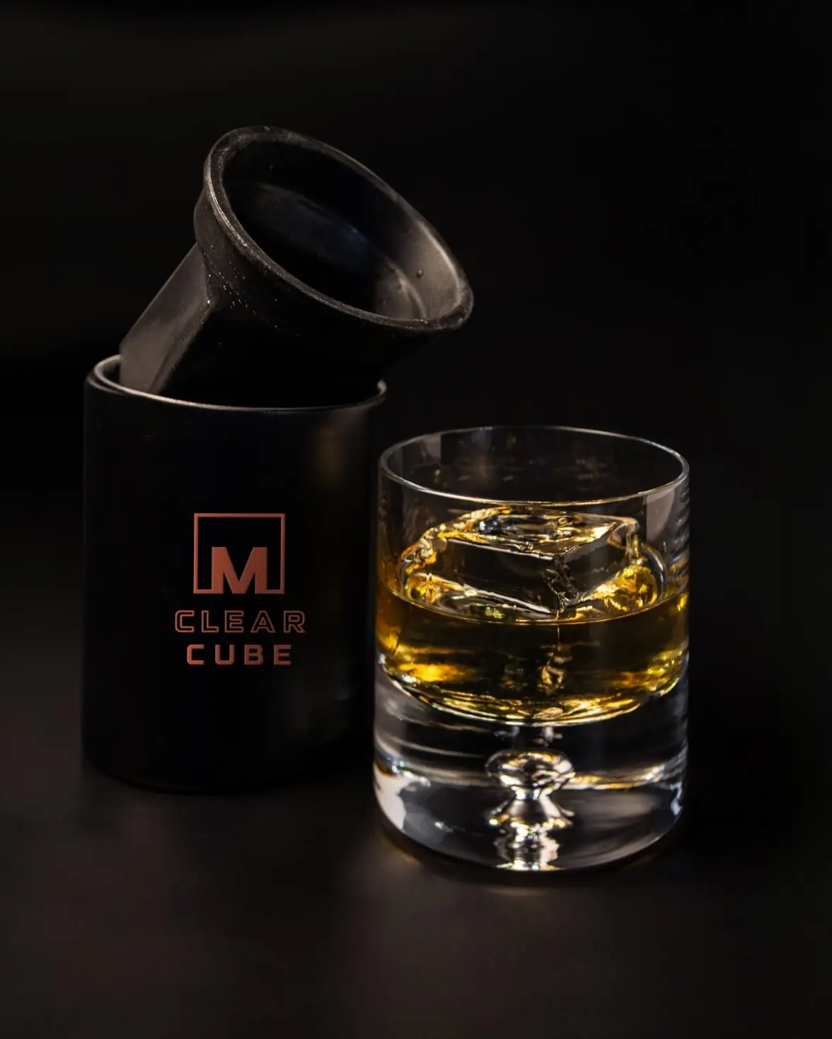 A glass of whiskey with a large clear ice cube sits next to a black cylindrical clear ice maker labeled M CLEAR CUBE on a dark background.