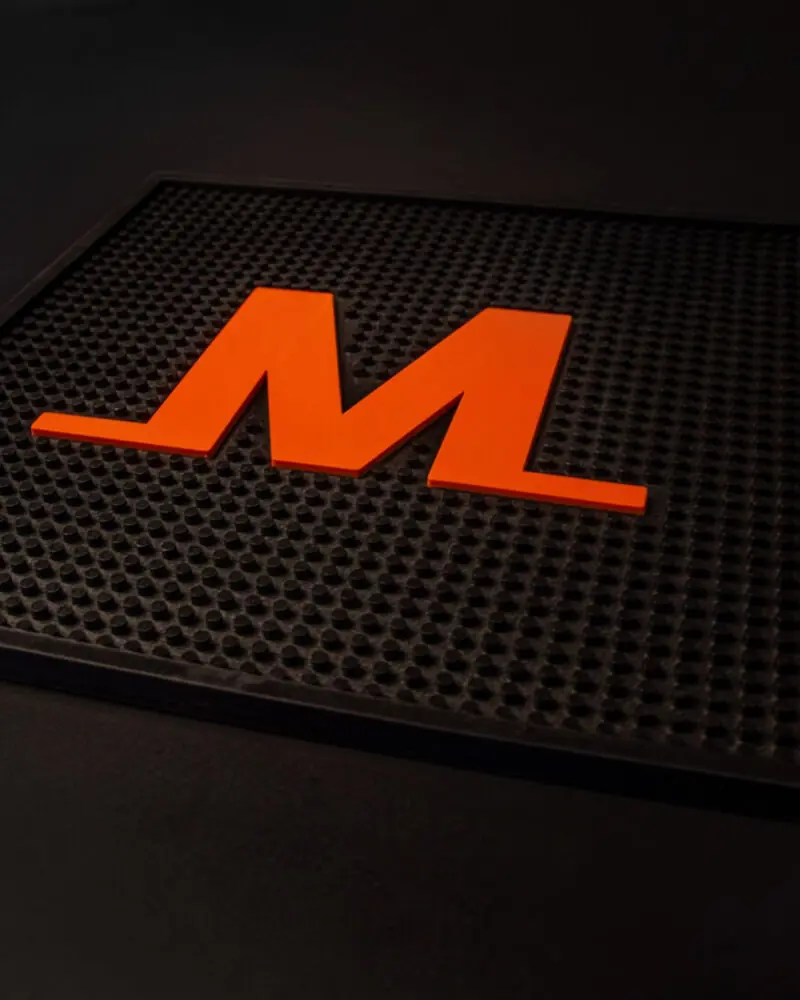 A red, stylized letter M sits on a black, textured rubber mat against a dark background.