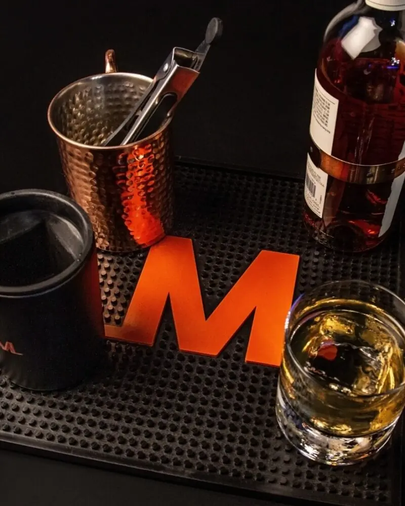 A glass of whiskey, an empty black cup, a copper mug with ice tongs, and a bottle of liquor sit on a bar mat with a large orange M on a black background.