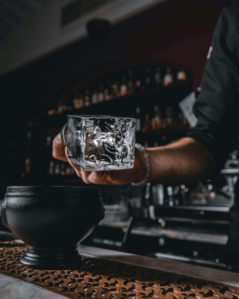 A bartender wearing a black shirt holds a textured, clear glass with a large ice cube above a bar counter with a black cup nearby. Shelves with bottles are visible in the dimly lit background.