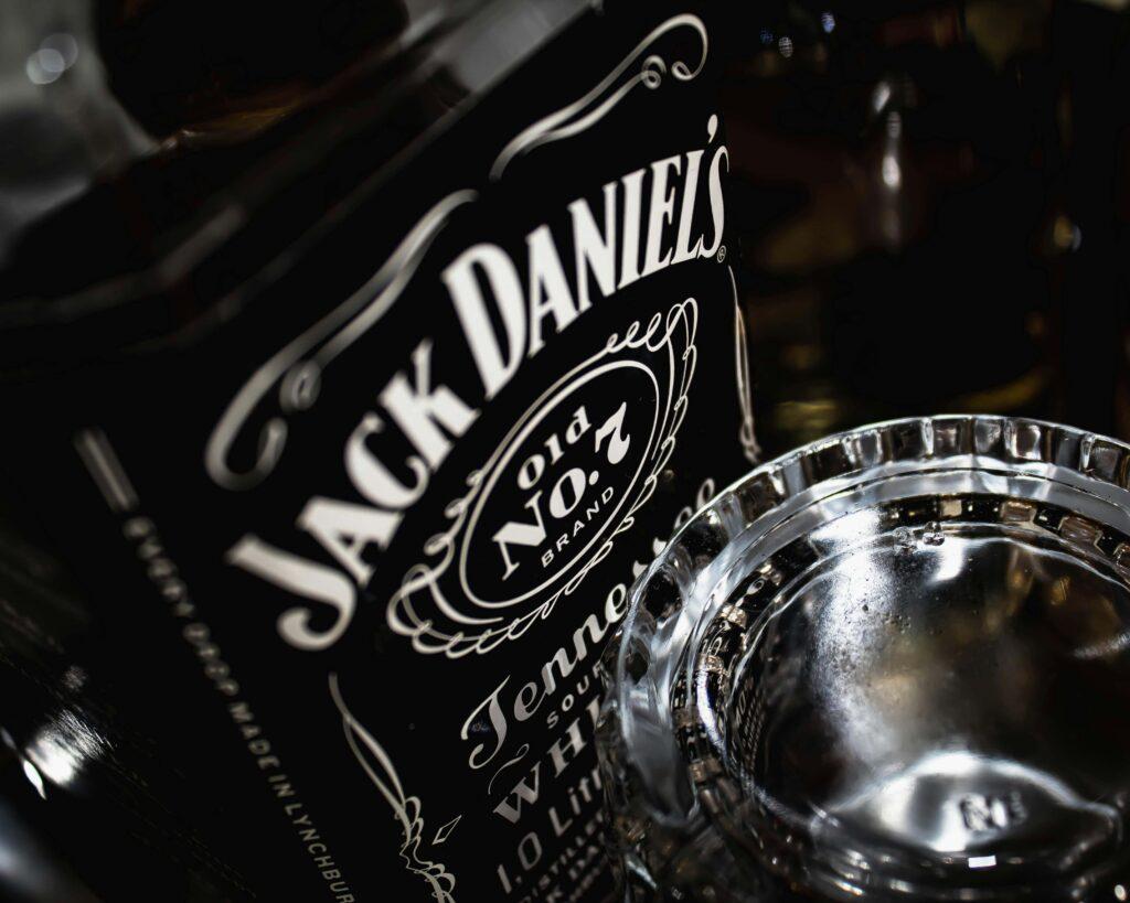 Close-up of a Jack Daniel’s Tennessee Whiskey bottle, an iconic American made liquor, partially obscured by a clear, empty glass ashtray in the foreground. The black and white label is prominently displayed.