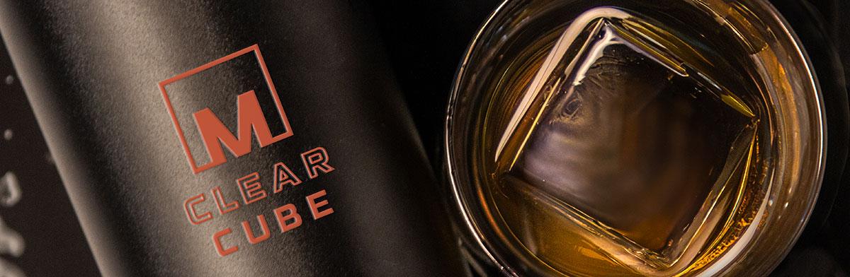 A close-up of a glass of whiskey with a large clear ice cube, next to a black container labeled M Clear Cube in orange lettering.