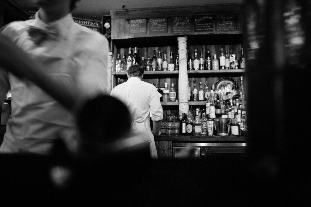A black and white image of a bar scene showing a bartender facing shelves full of bottles, with another person in the foreground, slightly blurred, wearing a white shirt and bow tie, capturing the artistry behind crafting the perfect cocktail.