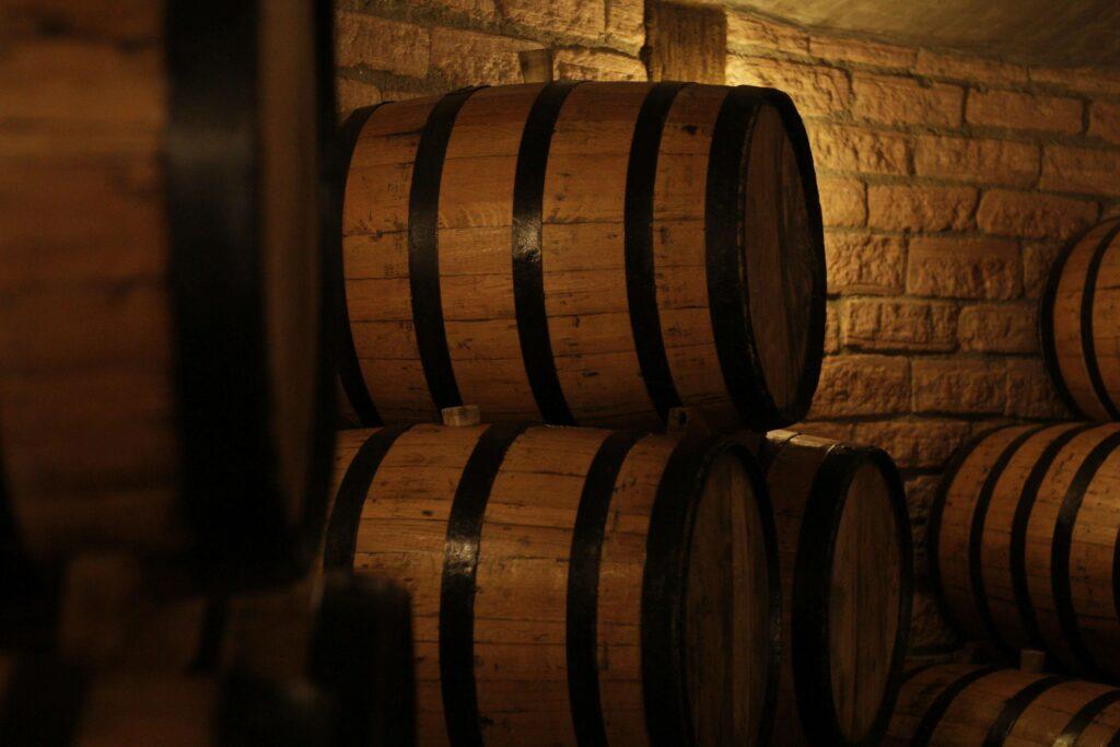 Wooden barrels with metal bands, essential for aging fine spirits, are stacked in a dimly lit cellar with brick walls, creating a rustic and cozy atmosphere.