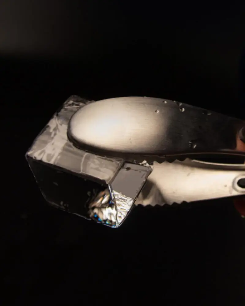A metal ice tong gripping a transparent, rectangular ice cube against a dark background, with some water droplets visible on the tong.