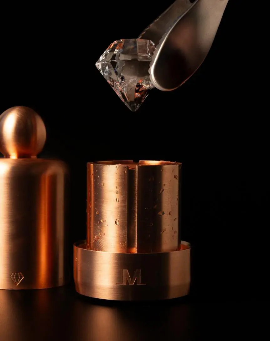 A Meltdown Mogul copper clear ice ball press sits on a dark surface as a large, diamond-shaped ice cube is held above it by metal tongs. The press and its components have a shiny, bronze finish.