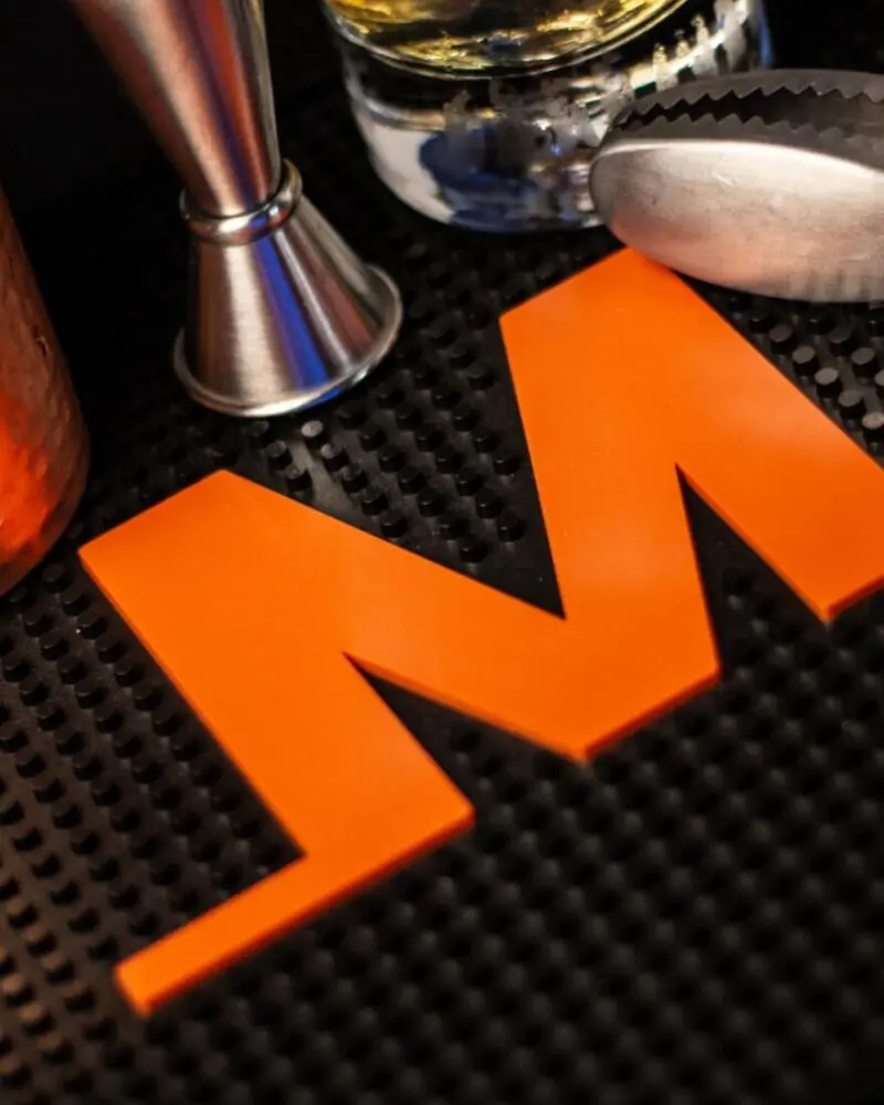 A close-up of a bright orange letter M on a black textured bar mat, with metal bar tools and a glass with a light-colored beverage nearby.