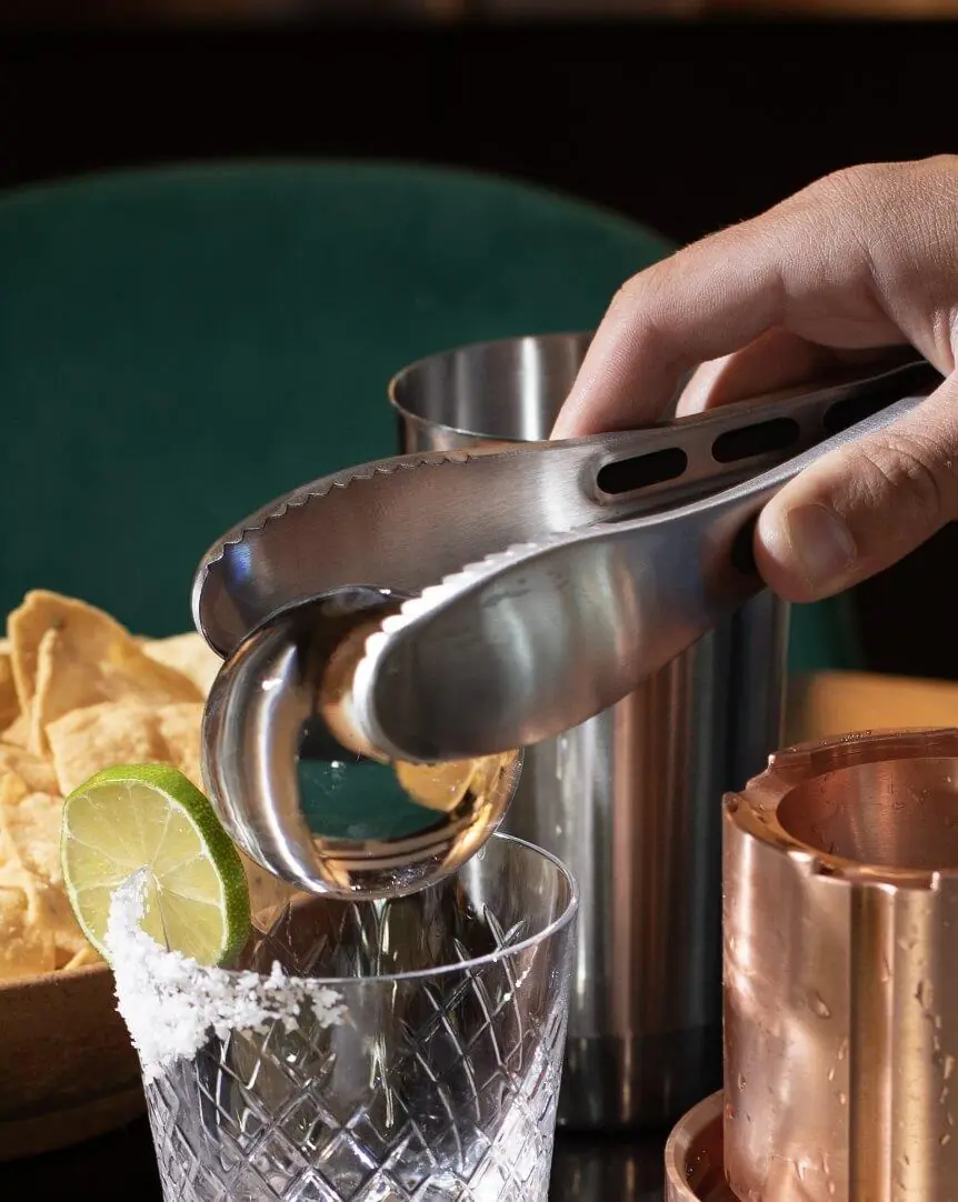 A hand uses metal tongs to place a large ice sphere into a glass rimmed with salt and garnished with a lime slice. A copper cup, a metal shaker, and a bowl of tortilla chips are nearby.