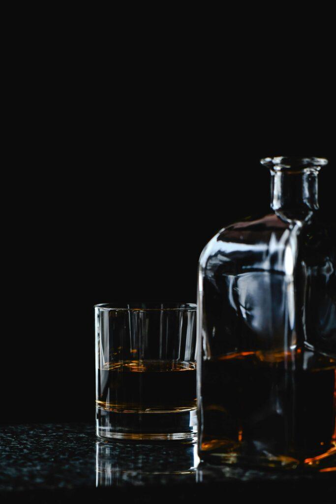 A glass of bourbon sits next to a partially filled whiskey decanter on a dark, reflective surface with a black background.