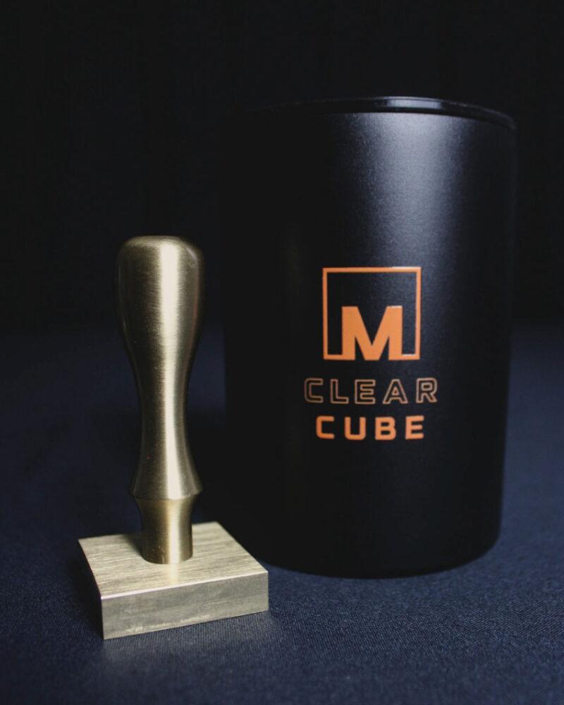 A brass stamp with a square base stands beside a black cylindrical container labeled M CLEAR CUBE in orange, both part of the Meltdown Clear Cube Ice Stamp Bundle, set against a dark background.