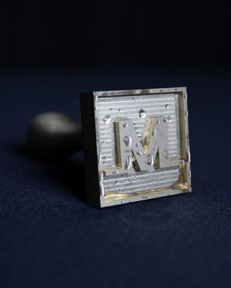 A metallic stamp with the letter M appears on a dark blue surface with water droplets—a perfect match for the Meltdown Clear Cube Ice Stamp Bundle.