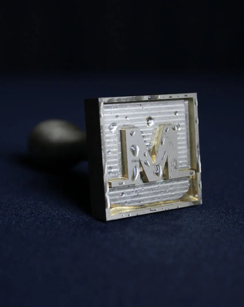 A metallic stamp with the letter M appears on a dark blue surface with water droplets—a perfect match for the Meltdown Clear Cube Ice Stamp Bundle.