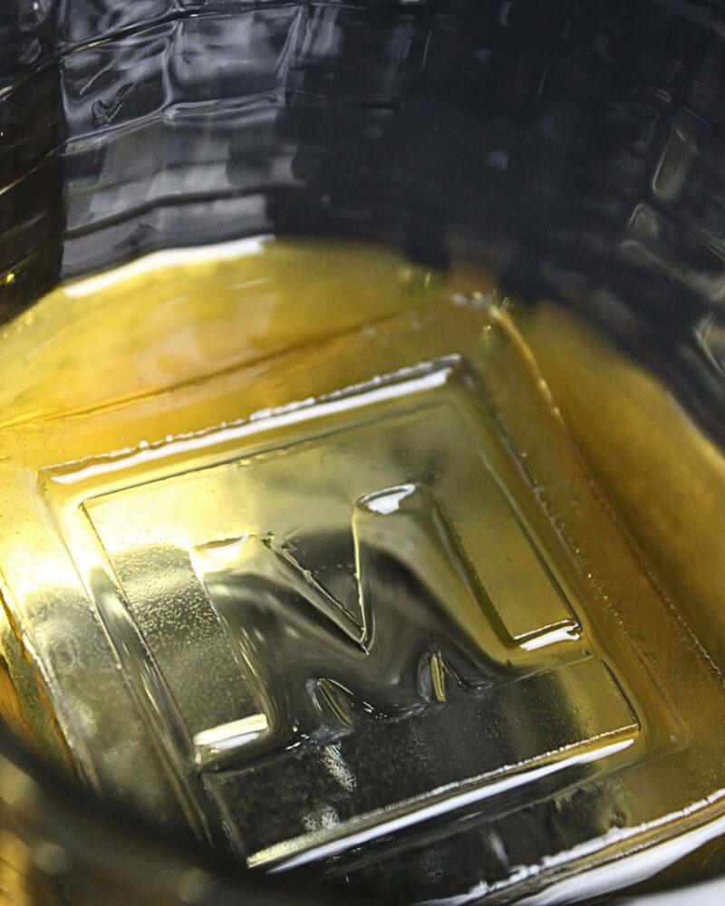 Close-up of a glass bottom with a raised M in the center, seen through golden liquid and enhanced by the clarity of the Meltdown Clear Cube Ice Stamp Bundle.