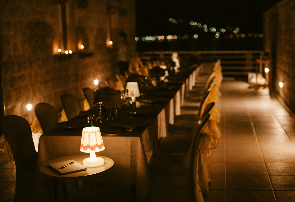 A long, elegant dining table set for an evening party is decorated with candles and lamps, surrounded by chairs on a terrace overlooking city lights at night. A small lit lamp and a guestbook sit on a side table in the foreground.